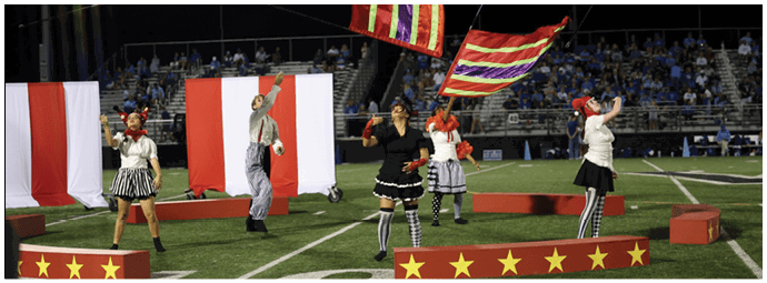 People dressed in colorful costumes perform a choreographed routine on a decorated stage set up on a sports field with an audience in the background. Descripci n generada con IA