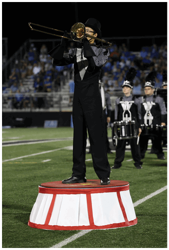 A person in a marching band uniform is playing a trombone on a football field with other band members and a crowd in the background. Descripci n generada con IA