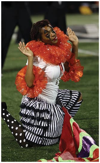 A person dressed in a colorful clown costume with a large red ruffled collar and striped skirt is kneeling on grass with hands raised near their face. Descripci n generada con IA