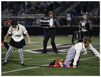 People in marching band uniforms and theatrical costumes perform on a football field with drums and colorful flags. Descripci n generada con IA