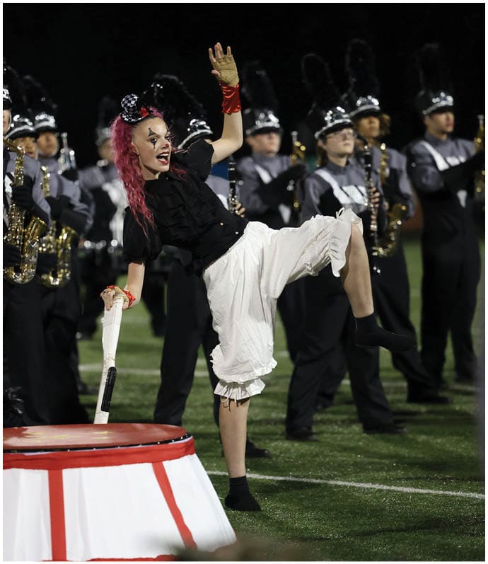 A person with bright pink hair and theatrical makeup is energetically performing with a drumstick on a large drum while a marching band in uniform plays instruments in the background on a field. Descripci n generada con IA