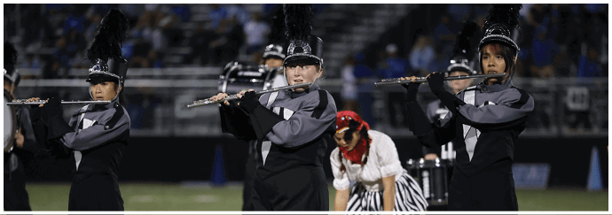 Three people in marching band uniforms are playing flutes on a field with a person in a red hat and striped pants in the background. Descripci n generada con IA