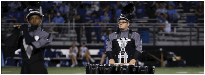 A person in a marching band uniform is playing a set of tenor drums on a football field with other band members and spectators in the background. Descripci n generada con IA