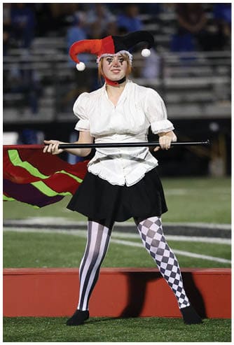 A person wearing a red and black jester hat, a white blouse, a black skirt, and mismatched black and white striped and checkered tights is holding a flag on a sports field. Descripci n generada con IA