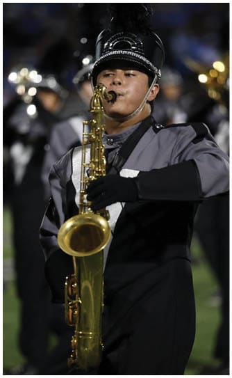 A person in a marching band uniform is playing a saxophone outdoors at night with other band members visible in the background. Descripci n generada con IA