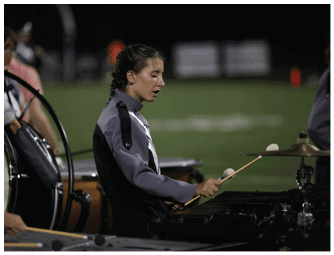 A person in a marching band uniform is playing a percussion instrument with mallets on a field at night. Descripci n generada con IA