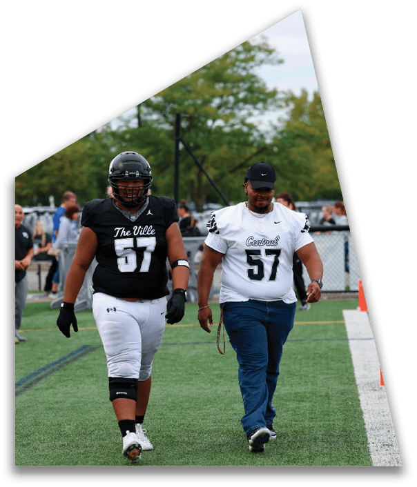 Two adults wearing football jerseys with the number 57 walk side by side on a sports field with trees and spectators in the background. Descripci n generada con IA