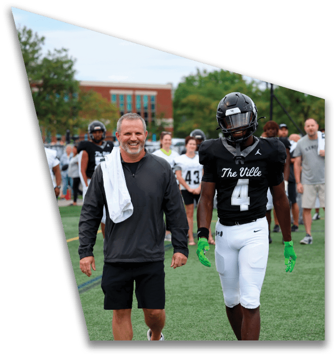 A person wearing a black sports jacket with a white towel over their shoulder walks beside a person in a black football uniform with the number four and green gloves on a sports field with other people and trees in the background. Descripci n generada con IA