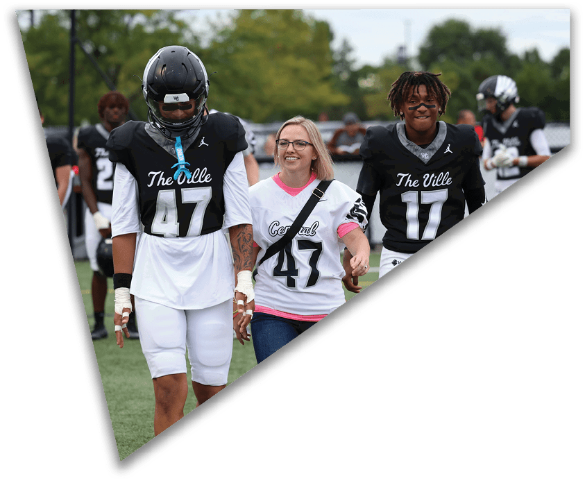 Two people in football uniforms walk on a field alongside a person in a casual sports jersey, all smiling and appearing to enjoy the moment. Descripci n generada con IA