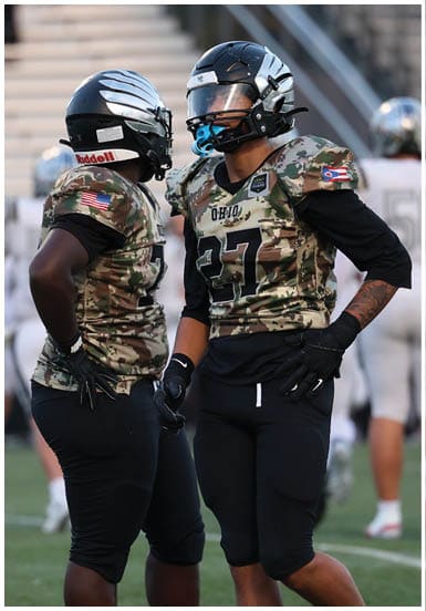 Two people wearing camouflage football uniforms and helmets are standing and talking on a football field with blurred players and spectators in the background. Descripci n generada con IA