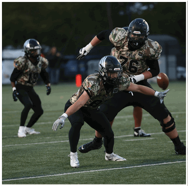Three people in camouflage football uniforms and helmets are actively playing on a grassy field with one person reaching out toward a football in midair. Descripci n generada con IA