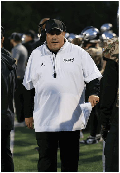 A person wearing a white sports shirt with “HAWKS" written on it and a black cap is standing on a sports field holding a piece of paper. Descripci n generada con IA