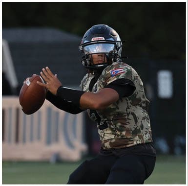 A person wearing a camouflage sports jersey and helmet is poised to throw a football on a grassy field during a game. Descripci n generada con IA