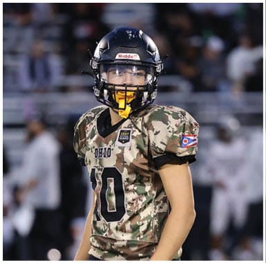 Two people wearing camouflage football uniforms and helmets are on a field with blurred spectators in the background. Descripci n generada con IA