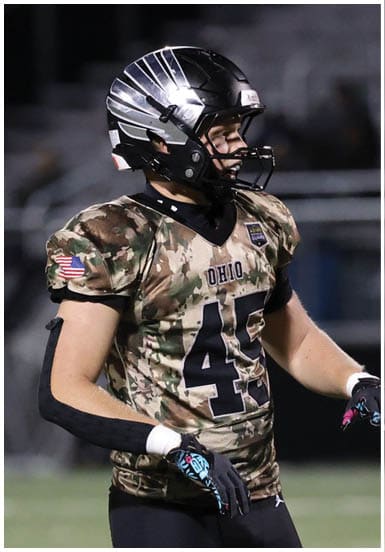 A person wearing a camouflage football jersey with the number 45 and a black helmet is standing on a football field. Descripci n generada con IA