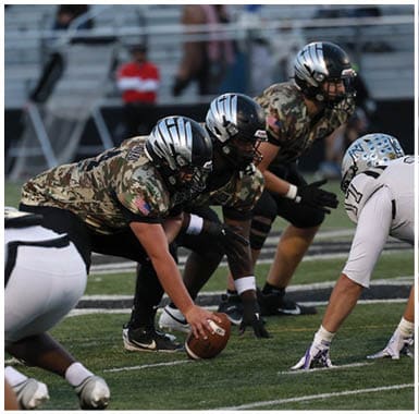 Players in camouflage and black helmets prepare to snap the football while opposing players in white uniforms line up on a football field. Descripci n generada con IA