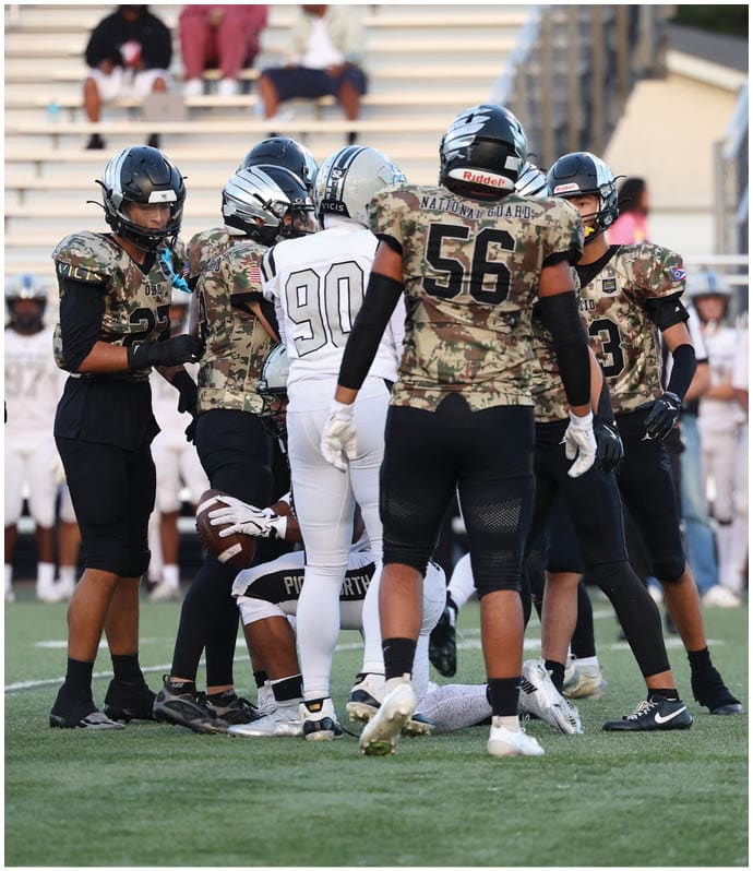 Players in football uniforms are gathered on a field with spectators and team members standing in the background near bleachers. Descripci n generada con IA