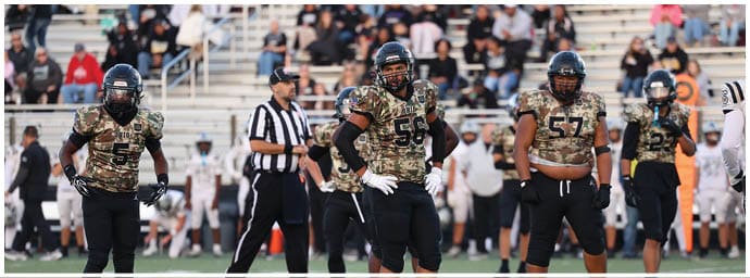 Four people in camouflage football uniforms stand on a field with a referee and spectators in the background. Descripci n generada con IA