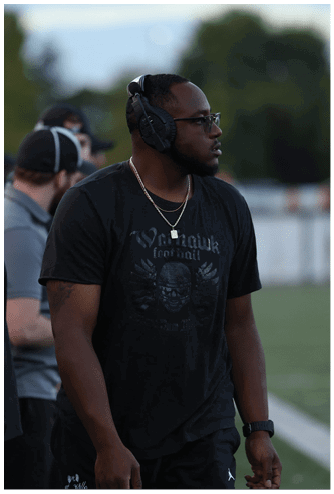 A person wearing glasses, headphones, a black graphic t-shirt, and a watch is standing on a sports field looking to the side. Descripci n generada con IA