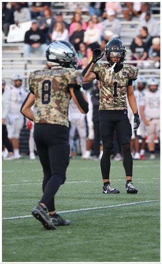 A person wearing a camouflage football jersey with the number one and a black helmet stands on a football field with a crowd and other players in white uniforms blurred in the background. Descripci n generada con IA