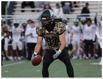 A person wearing a camouflage football jersey and black helmet is catching or holding a football on a field while other players and a referee are visible in the background. Descripci n generada con IA