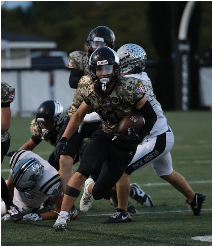 People wearing football uniforms are actively playing on a field with one person running while holding the ball and others attempting to block or tackle. Descripci n generada con IA