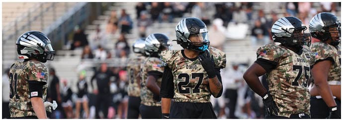People wearing camouflage football uniforms and helmets stand on a field during a game or practice. Descripci n generada con IA