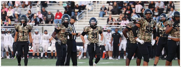 Several people in camouflage football uniforms stand on a field with a referee and other players in the background. Descripci n generada con IA