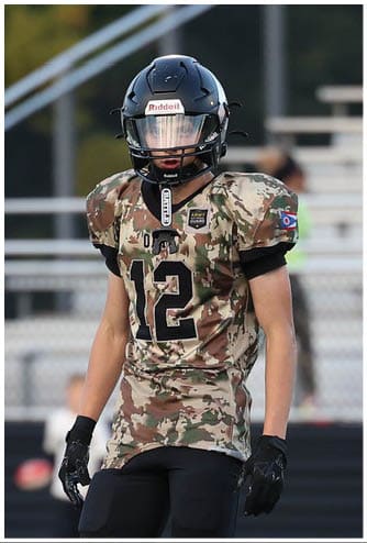 A person wearing a camouflage football jersey with the number 12, black pants, gloves, and a helmet is standing on a football field. Descripci n generada con IA