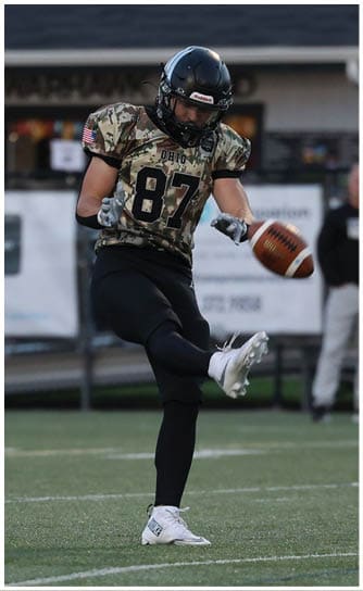 A person wearing a football uniform with the number 87 is kicking a football on a grassy field with a building and spectators in the background. Descripci n generada con IA