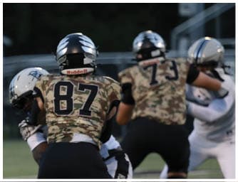 A person wearing a camouflage football uniform with the number 56 and a black helmet is on a football field with other players in the background. Descripci n generada con IA