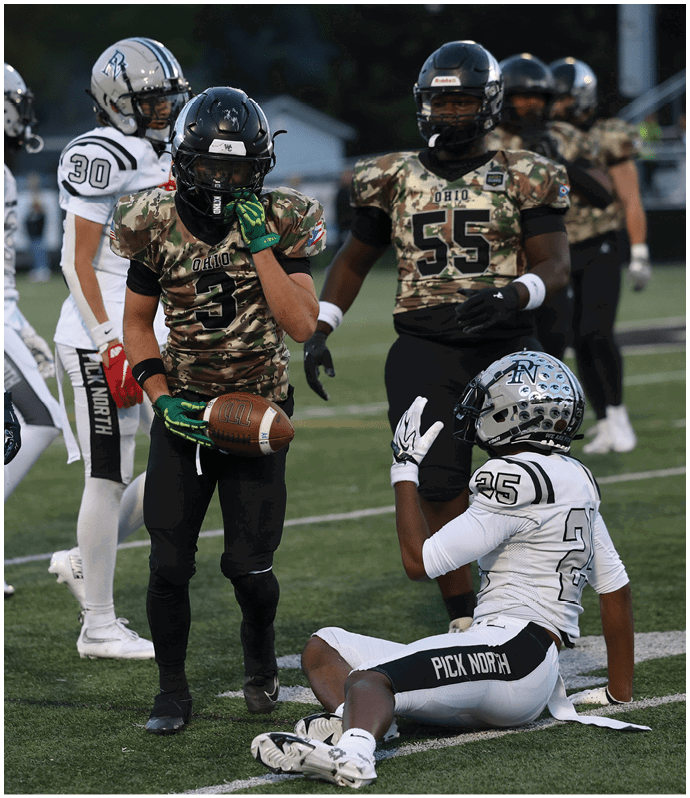 Four football players on a field are engaged in a game, with two wearing camouflage jerseys labeled “Ohio" and two in white jerseys, one holding a football. Descripci n generada con IA