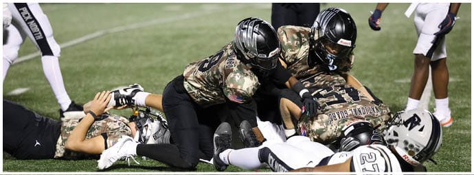 Two people in camouflage football uniforms and helmets are tackling a person lying on the ground during a game on a grassy field. Descripci n generada con IA