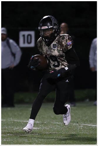 Two people in football uniforms are on a field at night with one running while holding the ball and the other lying on the ground, with several people standing in the background. Descripci n generada con IA