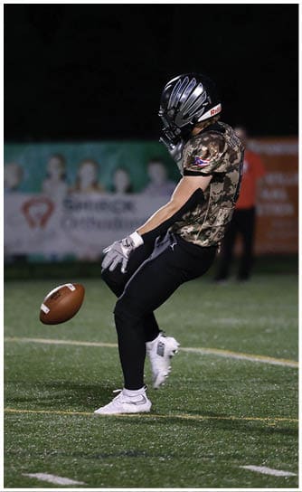 A person wearing a camouflage shirt, black pants, white cleats, gloves, and a black helmet is preparing to catch or kick a football on a grassy field at night. Descripci n generada con IA