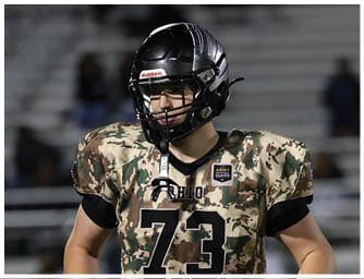 A person wearing a camouflage football jersey with the number 73 and a black helmet stands on a football field with another player in similar attire in the background. Descripci n generada con IA