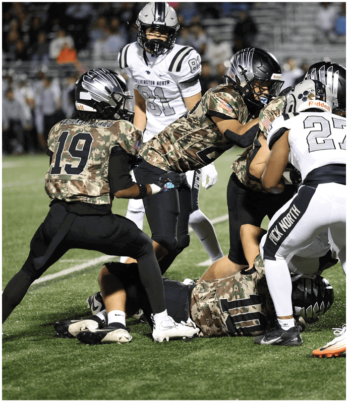 Several people wearing football uniforms are engaged in a play on a grassy field during a game. Descripci n generada con IA