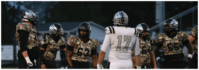 Adults wearing football uniforms in black and white are standing on a field, appearing to prepare for a play during a game. Descripci n generada con IA