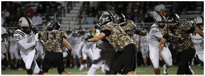 People wearing football helmets and uniforms are actively engaged in a play on a football field with a crowd in the background. Descripci n generada con IA
