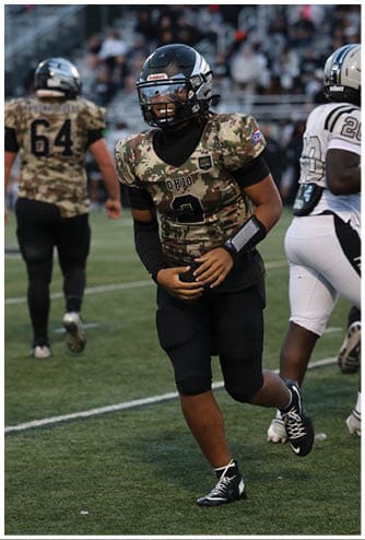 People wearing camouflage football uniforms are on a football field during a game, with one player running and others standing in the background. Descripci n generada con IA