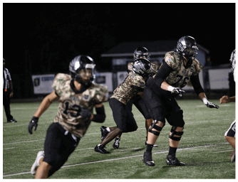 Several people wearing camouflage-patterned football uniforms and helmets are actively engaged in a nighttime football game on a field. Descripci n generada con IA