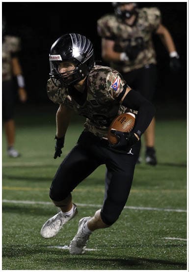 A person wearing a camouflage football uniform and helmet is running on a field while holding a football, with other players in similar uniforms visible in the background. Descripci n generada con IA