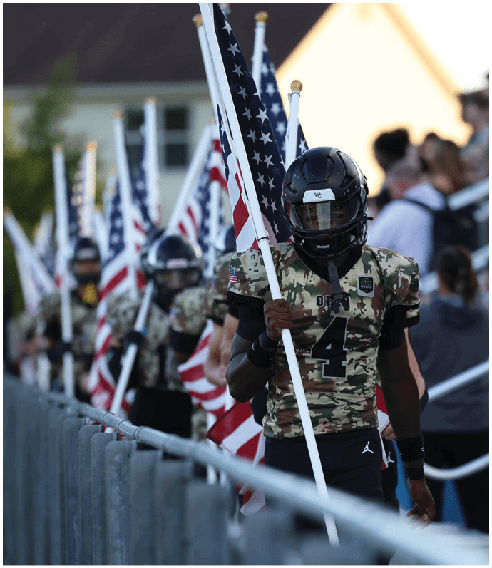 A line of people in camouflage football uniforms and helmets are holding American flags while walking along a fenced area with spectators in the background. Descripci n generada con IA
