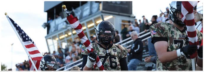 People in football uniforms holding American flags stand on a field with a crowd in the background. Descripci n generada con IA