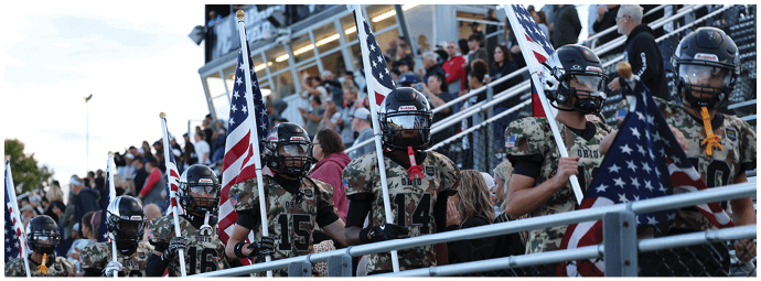 A group of people in football uniforms holding American flags stand behind a fence with a crowd seated in bleachers in the background. Descripci n generada con IA