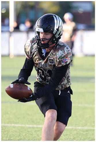 A person wearing a camouflage sports jersey and black helmet is holding a football and appears to be preparing to pass or run on a grassy field. Descripci n generada con IA
