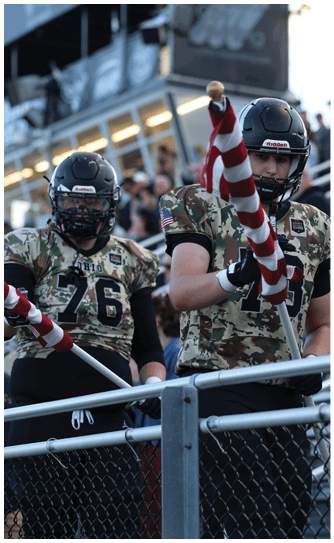 A person wearing a camouflage football uniform and helmet is holding a striped flag near a railing with people in the background. Descripci n generada con IA