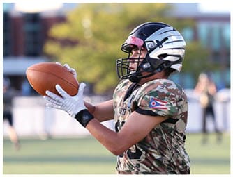 A person wearing a helmet and sports gear is standing on a grassy field with a blurred background of buildings and trees. Descripci n generada con IA