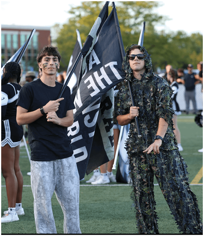 People on a sports field hold black and white flags, with one person wearing a camouflage outfit and another in a black shirt and light pants. Descripci n generada con IA