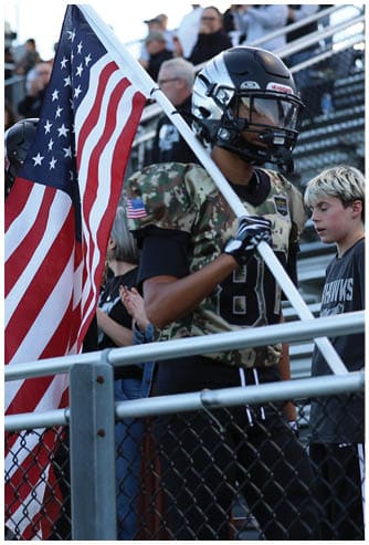 A person wearing a football helmet and camouflage jersey is holding an American flag while walking near a fence with other people in the background. Descripci n generada con IA